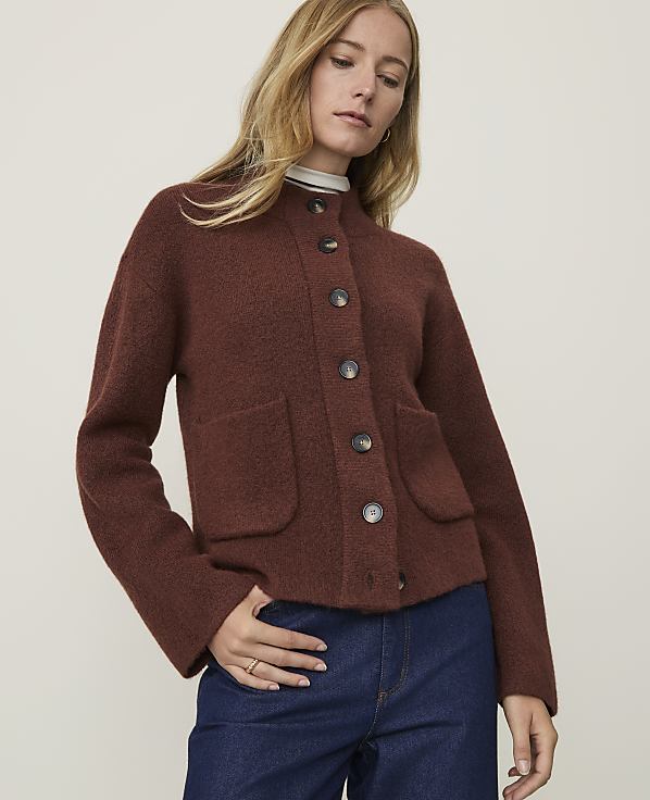 A woman in a brown cardigan and blue jeans stands against a plain, light-colored background.