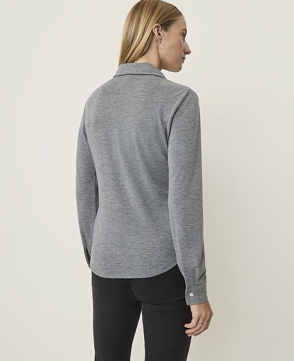 A woman in a grey long-sleeved shirt and black jeans stands against a plain, light-colored background.