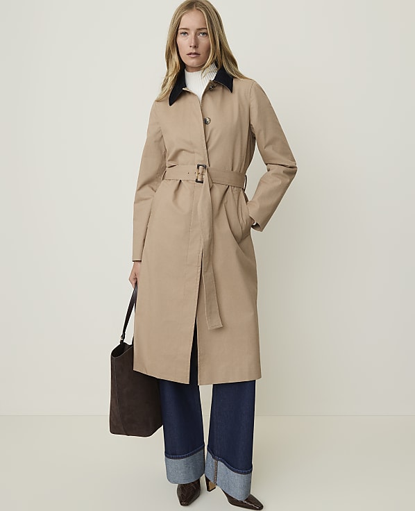 A woman in a tan trench coat and wide-leg jeans stands against a plain, light background, holding a dark tote bag.