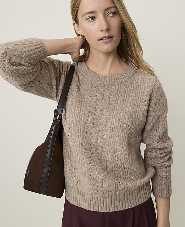 A woman in a cable-knit sweater and maroon skirt poses with a dark brown handbag against a plain, light background.