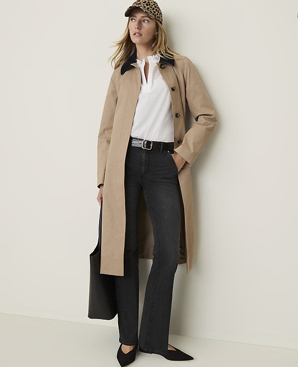 A woman in a tan trench coat, white blouse, dark jeans, and a leopard print cap stands against a plain, light-colored wall, holding a black tote bag.