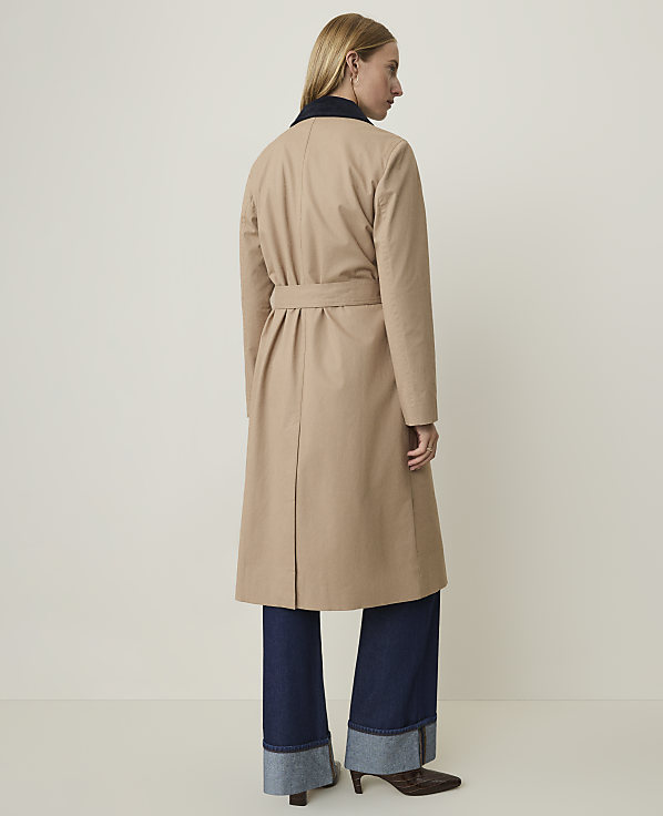 A woman in a tan trench coat and dark cuffed jeans stands against a plain, light background.