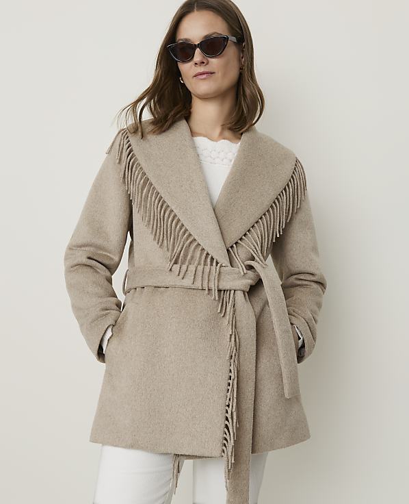 A woman in a fringed beige coat stands against a plain white background.