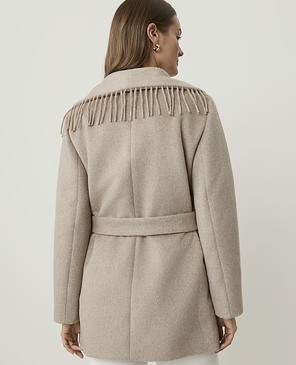 A woman in a beige fringed coat stands against a plain, light background.