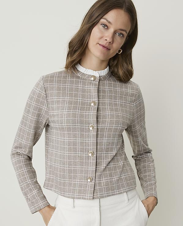 A woman in a plaid jacket and white pants stands against a plain background.