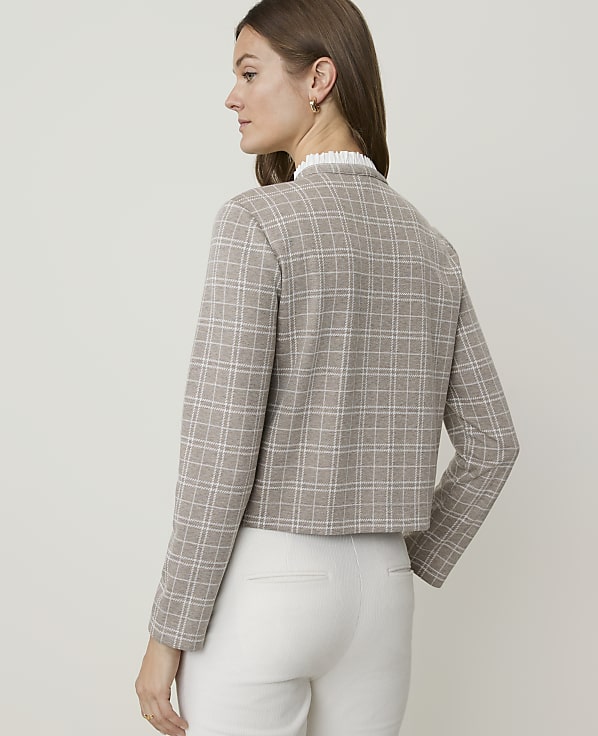 A woman in a plaid jacket and white pants stands against a plain background.