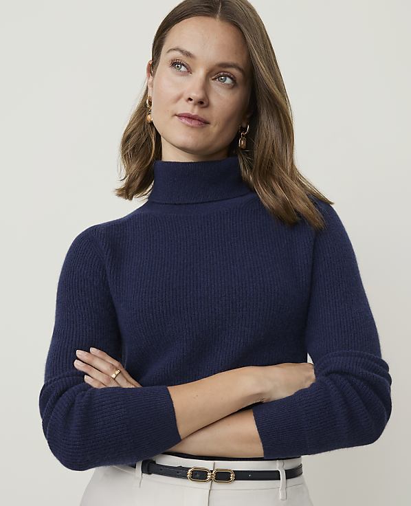 A woman in a navy turtleneck sweater and light-colored pants stands against a plain, light background.