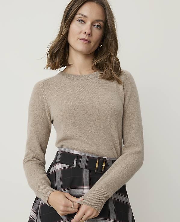 A woman in a beige sweater and plaid skirt stands against a plain background.