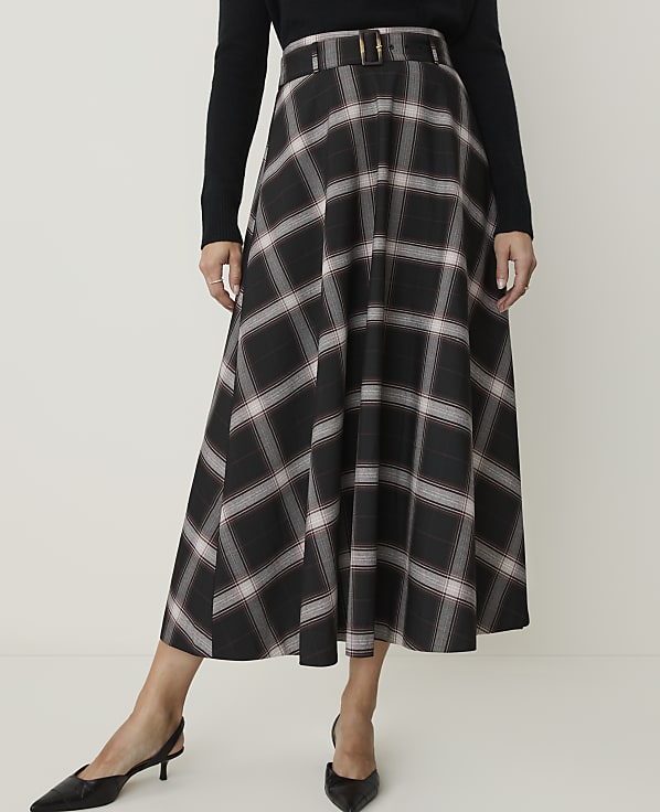A woman in a black long-sleeved top and a plaid midi skirt with a belted waist stands against a plain white background.