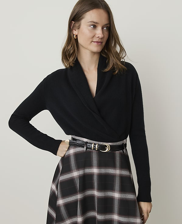 A woman in a black wrap sweater and plaid skirt stands against a plain, light background.