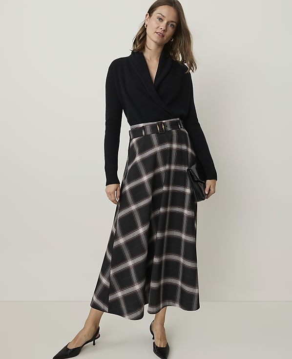 A woman in a black wrap sweater and a plaid midi skirt stands against a plain, light-colored background, holding a black clutch.