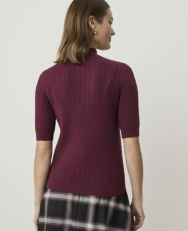 A woman in a burgundy cable-knit turtleneck sweater and plaid skirt stands against a plain, light-colored background.
