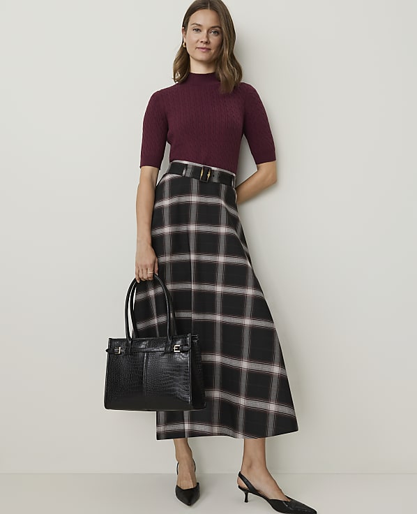 A woman in a burgundy mock-neck top and a plaid skirt stands holding a black crocodile-embossed tote bag against a plain, light-colored wall.