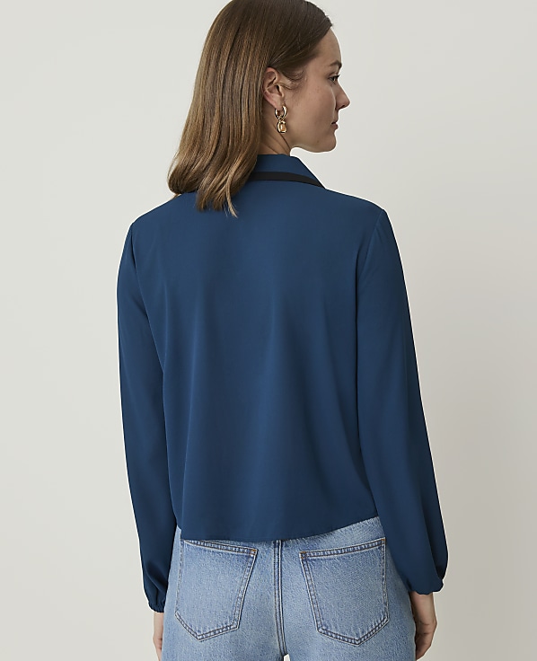 A woman in a teal blouse and jeans stands against a plain white background.
