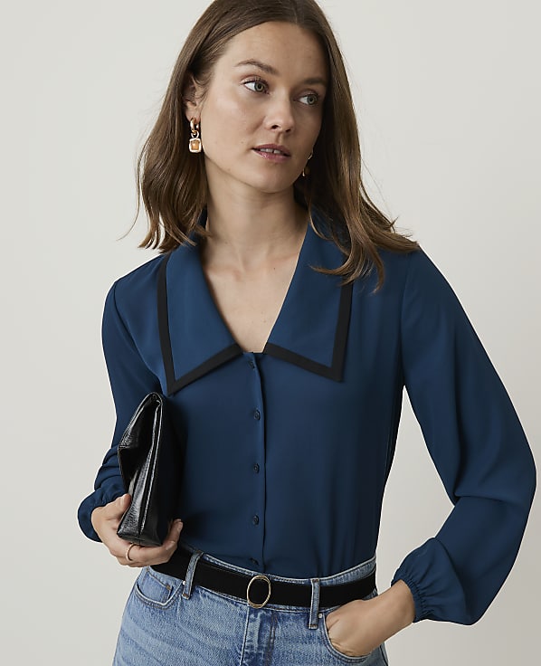 A woman in a teal blouse and jeans holds a black clutch in front of a plain background.