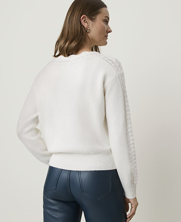 A woman in a white cable-knit sweater and blue leather pants stands against a plain white background.