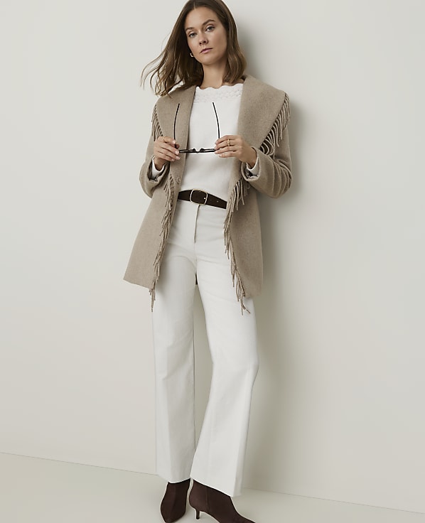 A woman in a fringed jacket and white pants stands against a plain, light-colored wall.