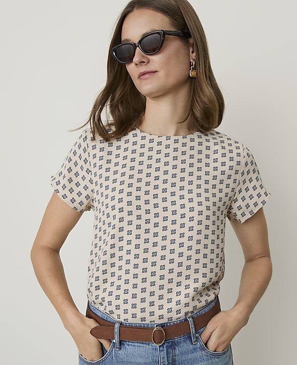 A woman in a patterned top and sunglasses stands against a plain background.