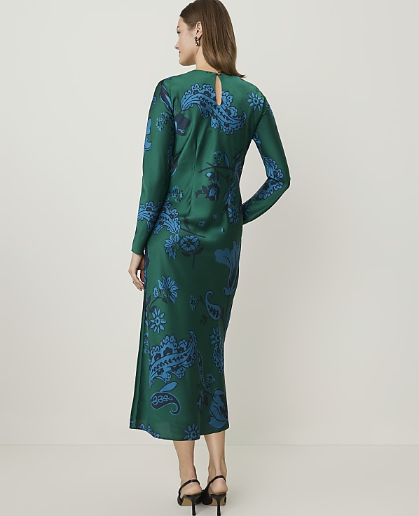 A woman in a green paisley-print dress stands against a plain, light-colored background.