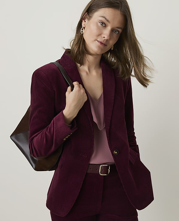 A woman in a burgundy corduroy suit with a pink camisole stands against a plain, light-colored background, carrying a brown tote bag.