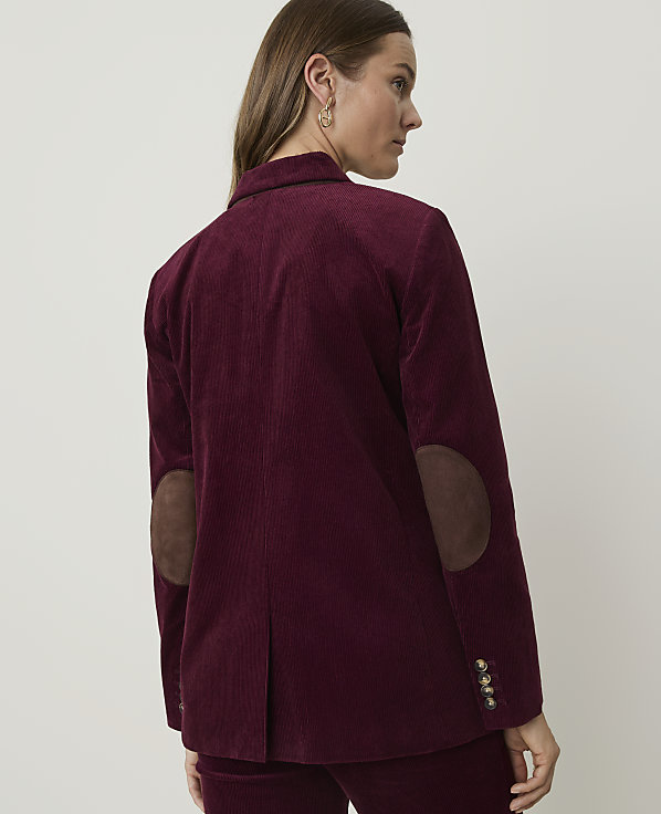 A woman in a burgundy corduroy blazer with suede elbow patches stands against a plain, light-colored background.