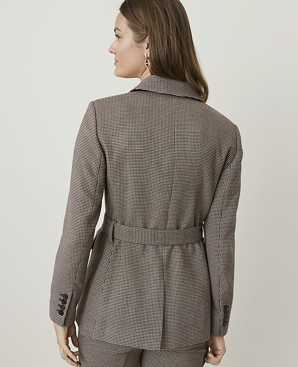 A woman in a brown checkered blazer and matching pants stands against a plain, light-colored background.