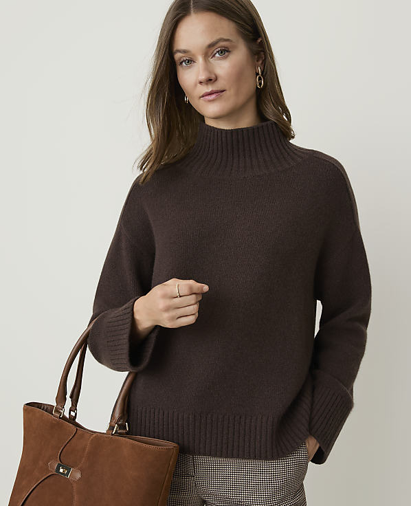 A woman in a brown turtleneck sweater and plaid pants holds a brown leather tote bag against a plain background.
