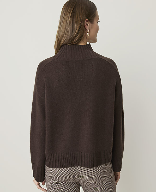 A woman in a brown turtleneck sweater and plaid pants stands against a plain, light gray background.