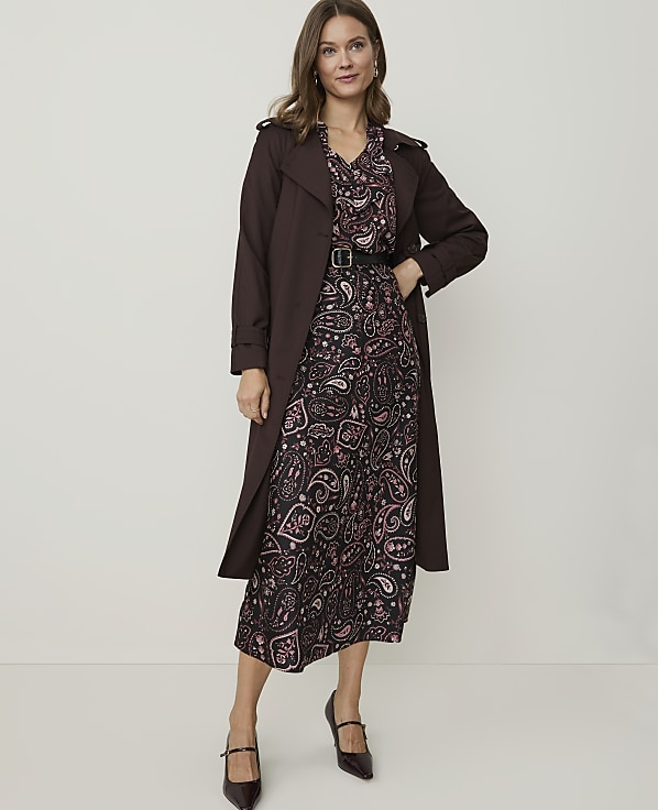 A woman in a patterned dress and brown trench coat stands against a plain white background.