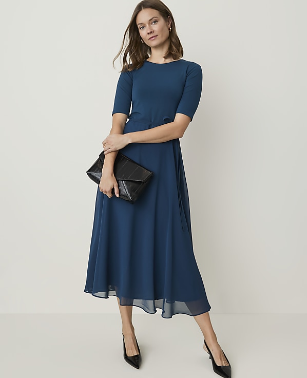 A woman in a teal dress and black heels holds a black clutch in front of a plain white background.