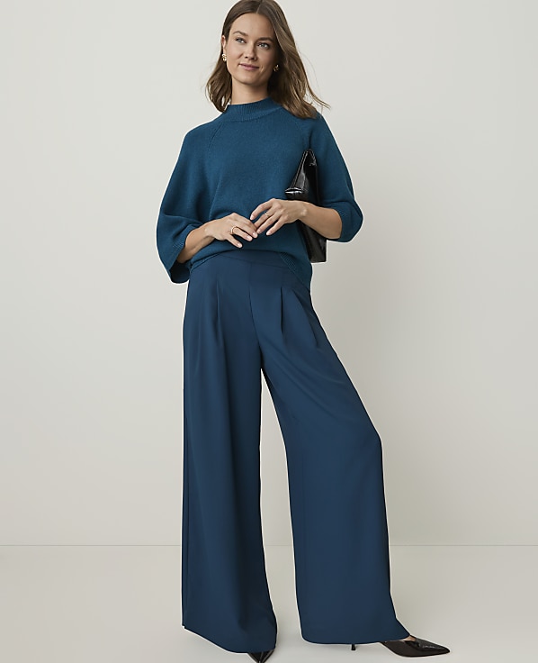 A woman in a teal sweater and wide-leg pants stands against a plain, light background, holding a black purse.