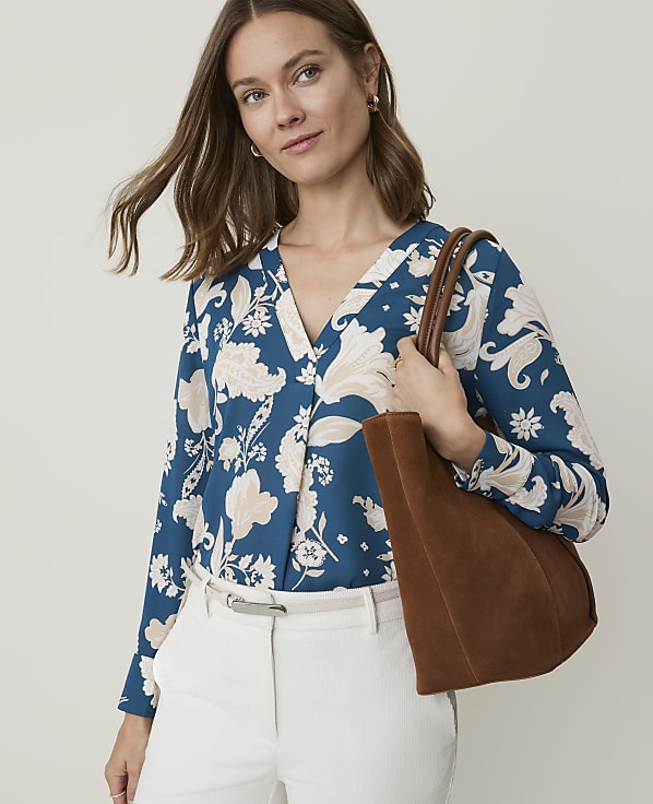 A woman in a floral blouse and white pants holds a brown suede tote bag against a plain background.