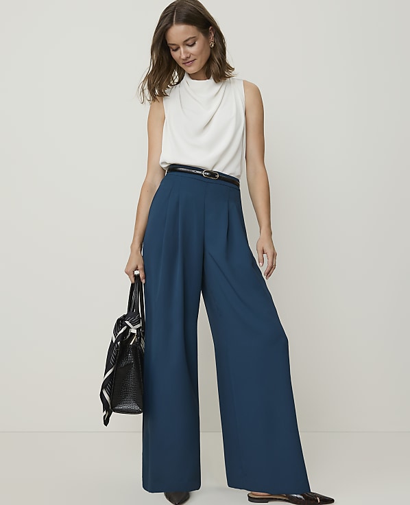 A woman in a white sleeveless top and teal wide-leg pants stands against a plain background, holding a black tote bag.