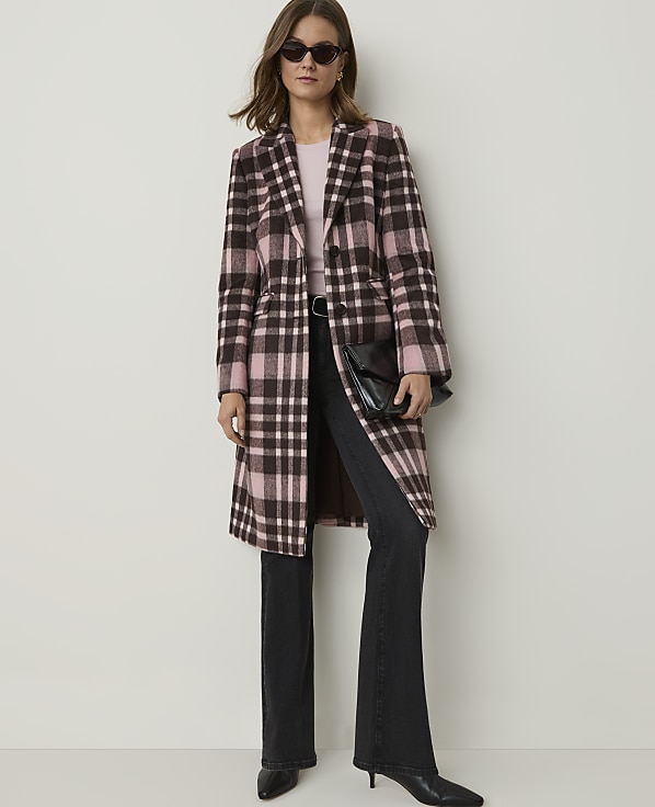 A woman in a pink and brown plaid coat and dark jeans stands against a plain, light-colored wall.