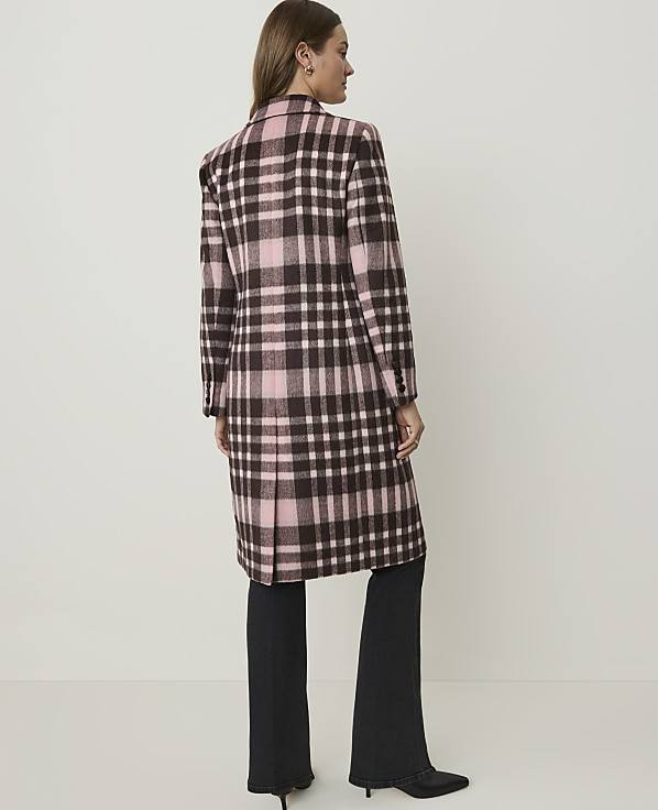 A woman in a pink and brown plaid coat stands against a plain, light-colored background.
