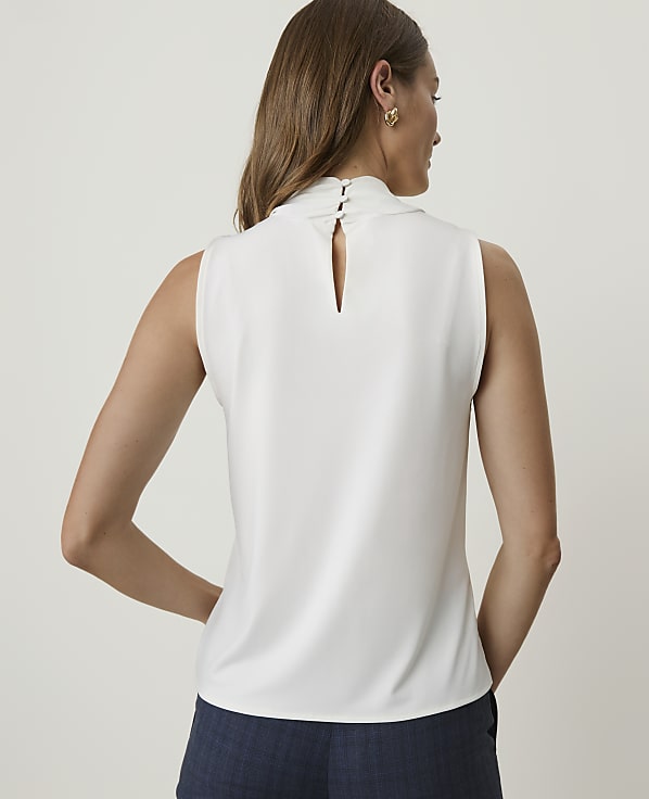 A woman in a white sleeveless top and blue shorts stands against a plain white background.