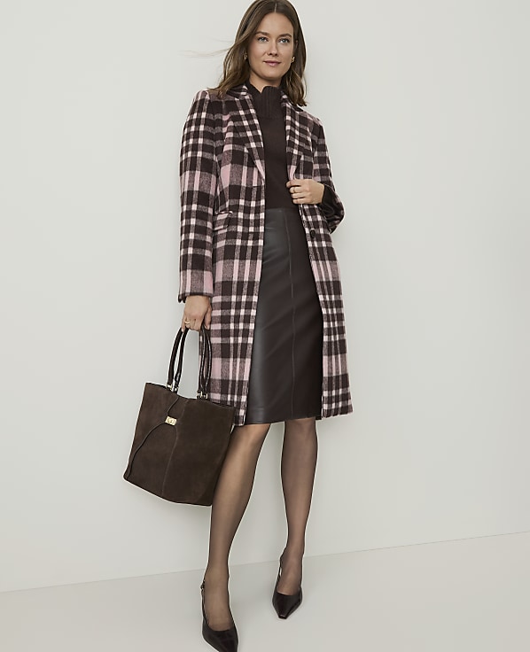 A woman in a plaid coat and leather skirt stands against a plain background, holding a brown suede tote bag.