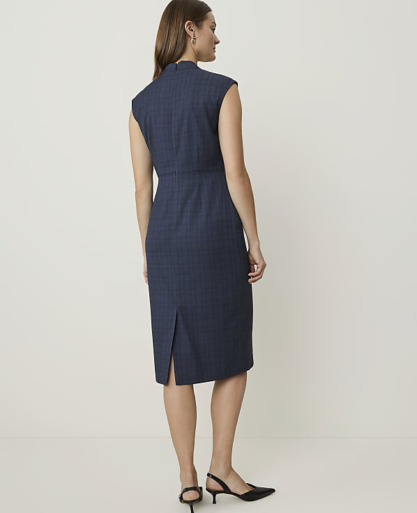 A woman in a blue plaid dress stands against a plain, light-colored background.