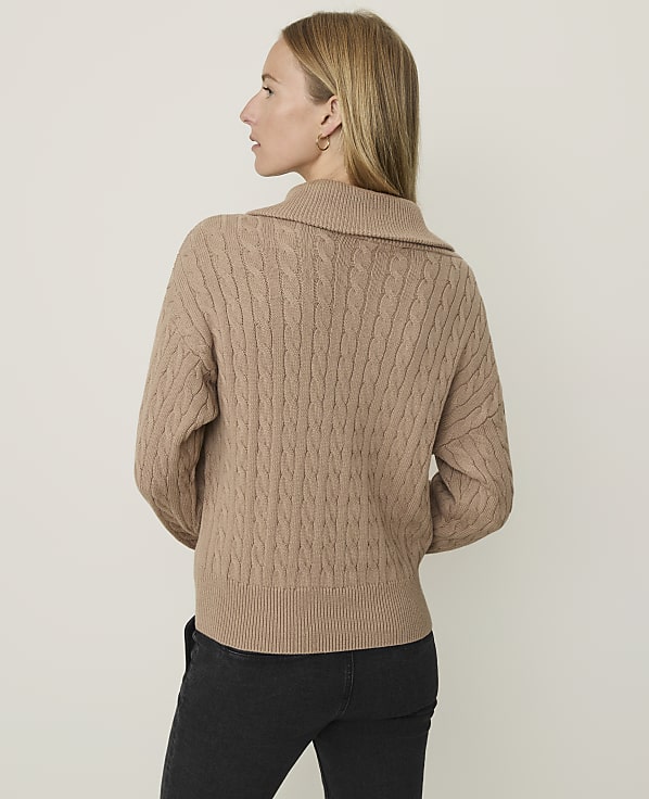 A woman in a cable-knit sweater stands against a plain, light-colored background.