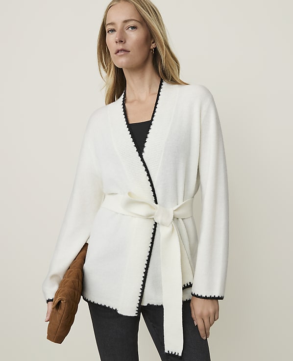 A woman in a white belted cardigan and dark pants holds a brown clutch in front of a plain background.