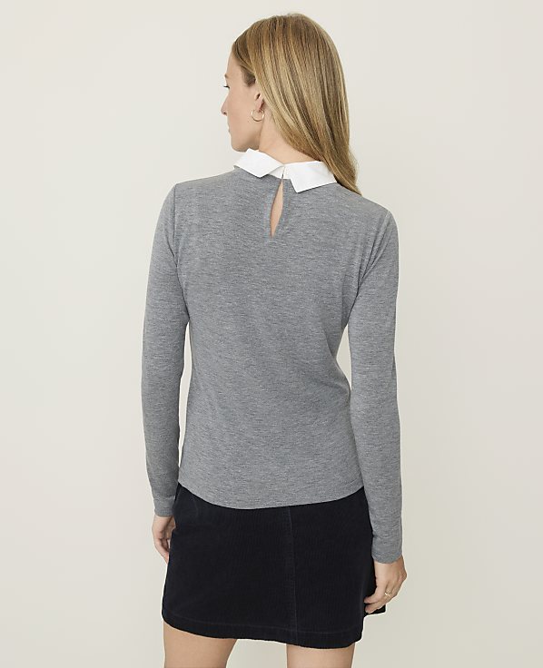 A woman in a gray sweater with a white collar and a dark skirt stands against a plain, light-colored background.