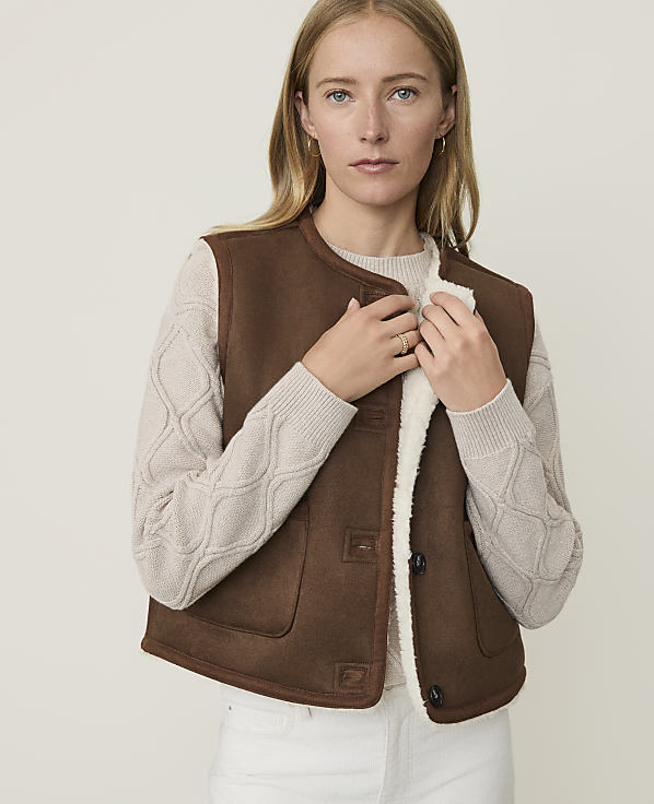 A woman in a brown shearling vest and cream sweater stands against a plain, light background.