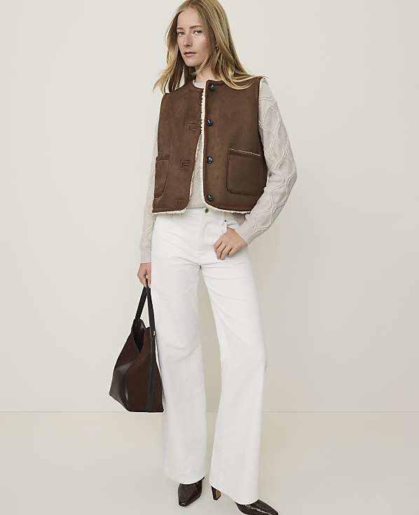 A woman in a brown vest and white pants stands against a plain, light background, holding a dark brown purse.