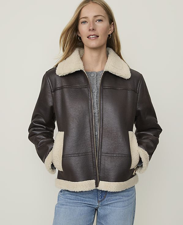 A woman in a brown faux-leather jacket with a cream shearling collar and cuffs stands against a plain, light background.