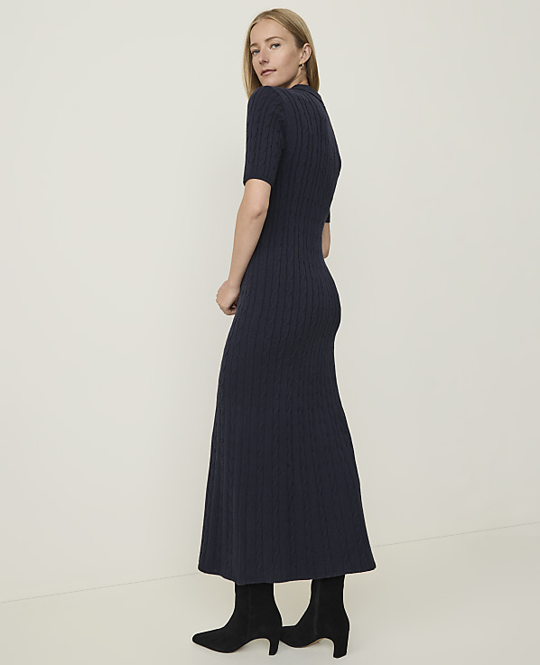 A woman in a navy cable-knit dress and black boots stands against a plain, light-colored background.