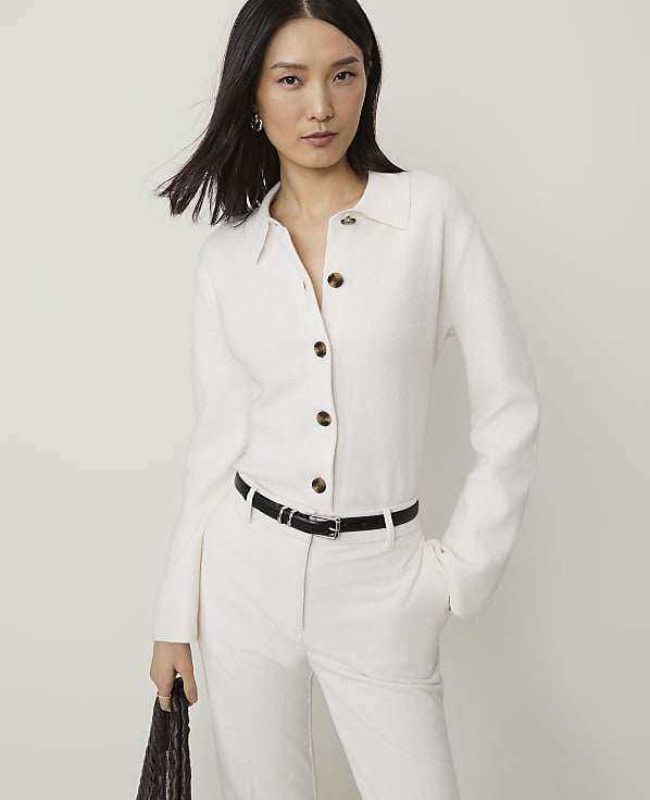A woman in a white cardigan and pants stands against a plain white background.