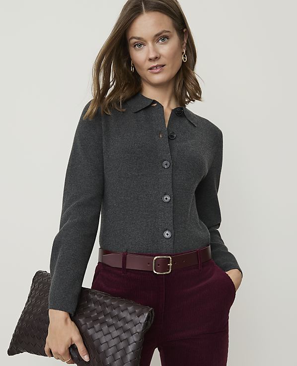 A woman in a dark grey collared shirt and burgundy pants holds a woven clutch in front of a plain background.