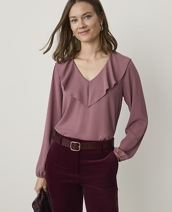 A woman in a mauve ruffled blouse and burgundy corduroy pants stands against a plain, light gray background.
