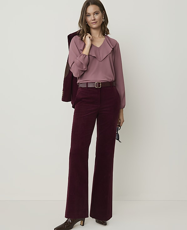 A woman in a mauve ruffled blouse and burgundy corduroy pants stands against a plain, light background, with a maroon jacket draped over her shoulder.