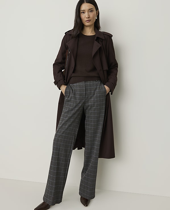 A woman in a brown trench coat and plaid pants stands against a plain white background.